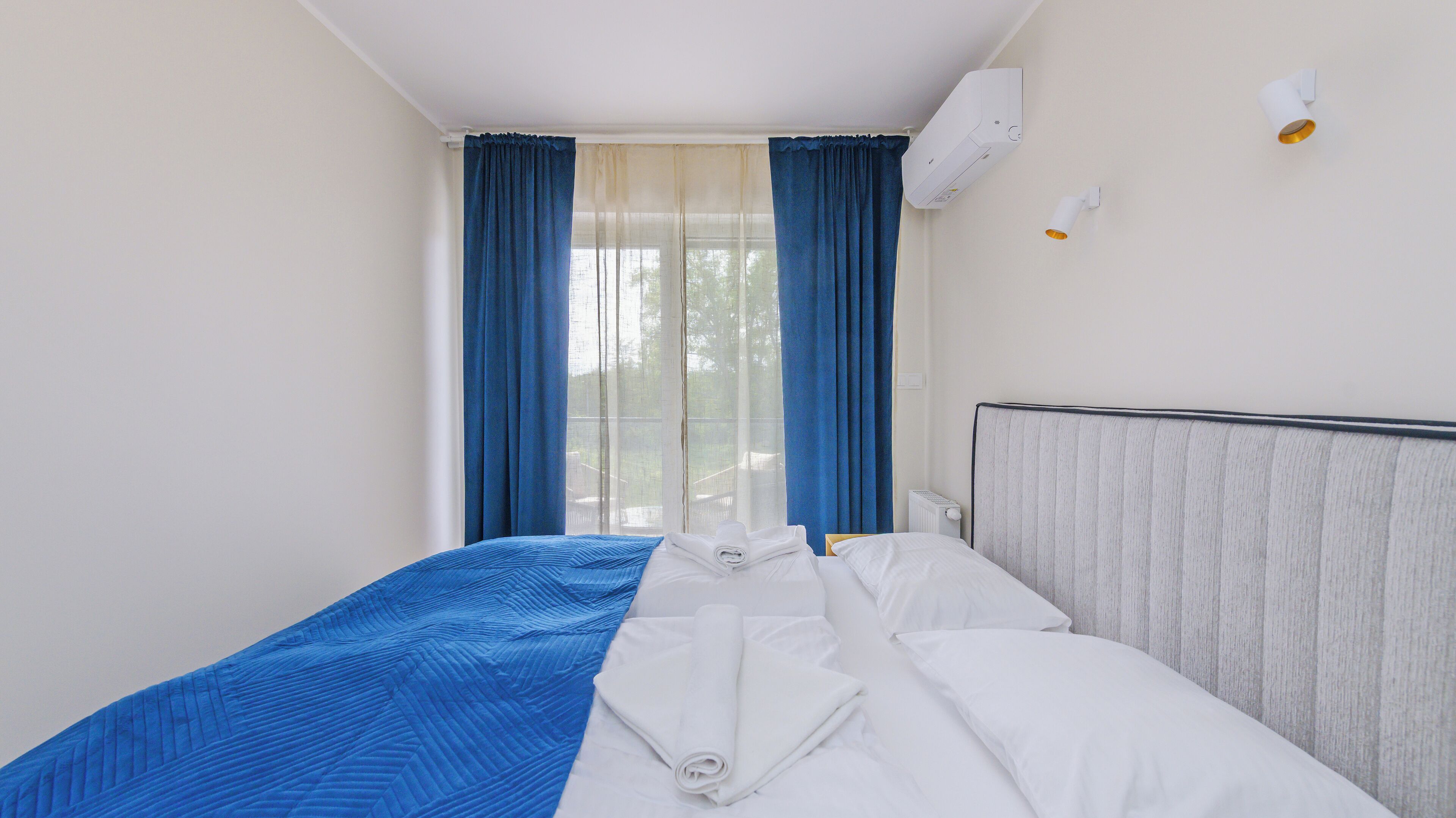 Une chambre d’hôtel avec un lit, une fenêtre avec des rideaux, un appareil de climatisation et un luminaire fixé au mur.