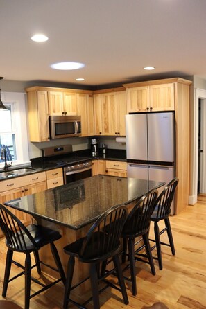 A modern kitchen with wooden cabinets, a stainless steel refrigerator, a microwave, and a large island with bar stools. - Private, River, Hot tub, Fire table, fishing (Marion)