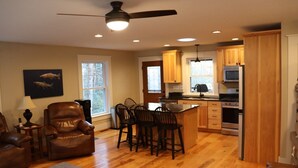 A spacious living room with a leather sofa, a dining area with chairs, a kitchen with wooden cabinets and appliances, and a ceiling fan.