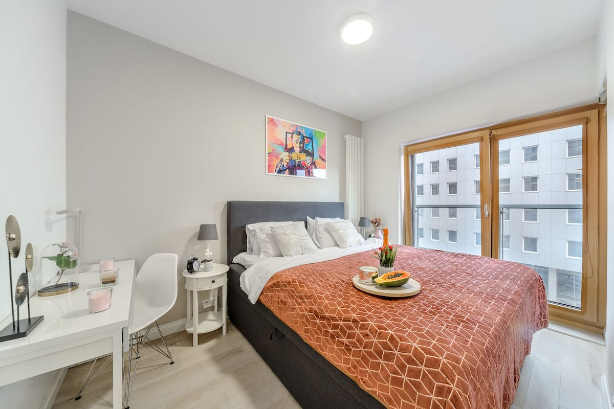 A modern bedroom with a large bed, a desk, a chair, and a window with a view of buildings.