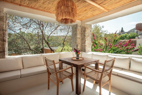 Zona pranzo con tavolo e sedie in legno, soffitto intrecciato e vista su alberi e fiori.