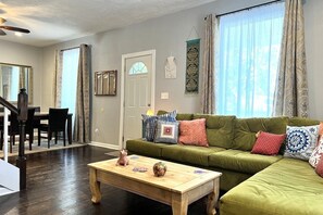 Un salon avec un canapé vert, une table basse en bois et des coussins décoratifs. On y trouve également un coin repas avec une table et des chaises, ainsi qu’un escalier menant à un autre niveau.