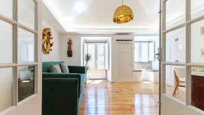 Un vaste espace de vie agrémenté d’un canapé vert, d’un luminaire suspendu doré et offrant une vue sur une cuisine, accessible par une porte ouverte.