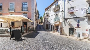 Une rue pavée bordée de bâtiments des deux côtés, avec des tables et des chaises installées à l’extérieur d’un café, et un panneau indiquant l’emplacement d’un distributeur automatique à proximité.