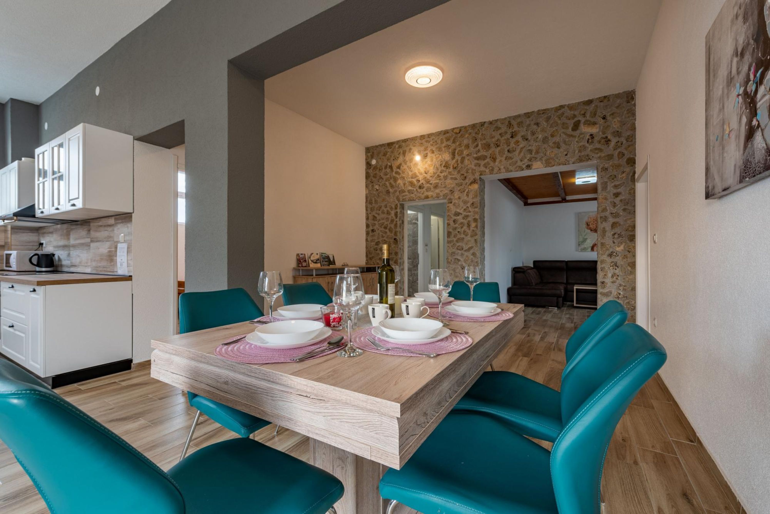 Een eetgedeelte met een houten tafel voor vier personen, teal stoelen en een keuken op de achtergrond.