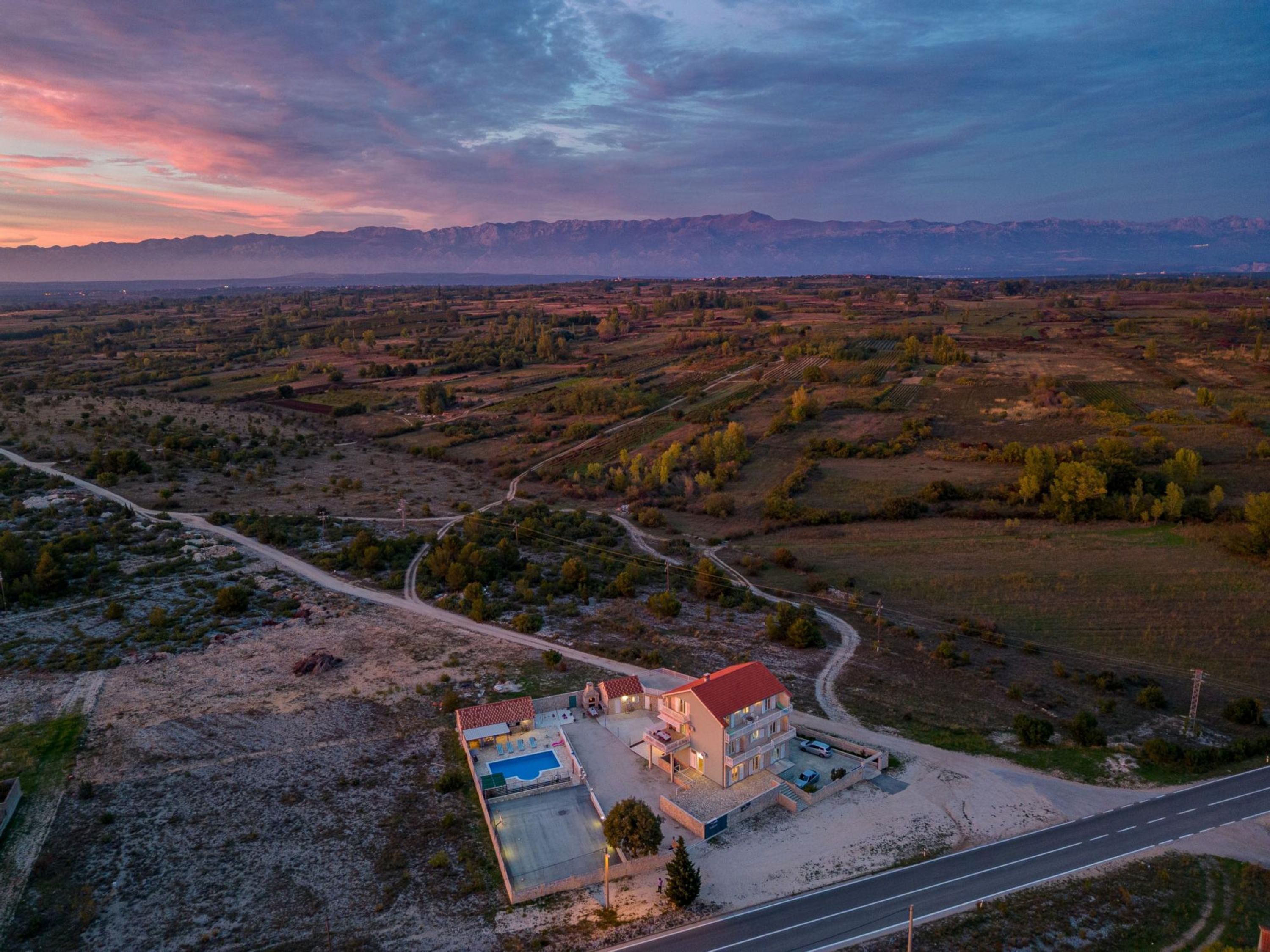 An aerial view of a two-story house with a swimming pool, surrounded by a rural landscape with fields and mountains in the background.