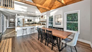 Een ruime keuken met een houten plafond, een eethoek met een grote tafel en stoelen, en uitzicht op bomen door grote ramen.