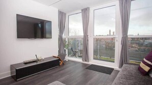 Una sala de estar moderna con un televisor de pantalla plana, un mueble negro para entretenimiento, un sofá gris y un amplio ventanal corredizo que da a un balcón exterior.