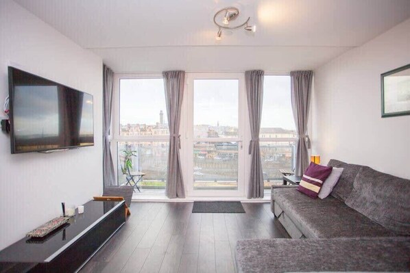 A modern living room with a flat-screen TV, a grey sofa, and a large window with curtains.