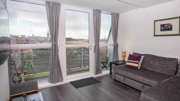 Una sala de estar moderna con un sofá gris, una mesita de noche con una lámpara y vistas de la ciudad a través de amplias ventanas con cortinas grises.