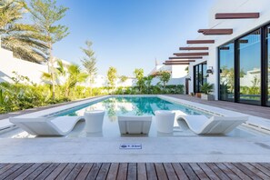 A modern pool area with lounge chairs and a clear blue pool, surrounded by greenery and a white building with large windows.