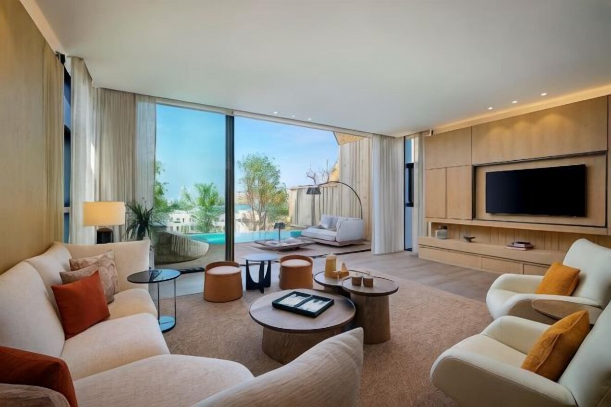 A spacious living room with a large flat-screen TV, comfortable seating, a coffee table, and a view of an outdoor pool area.