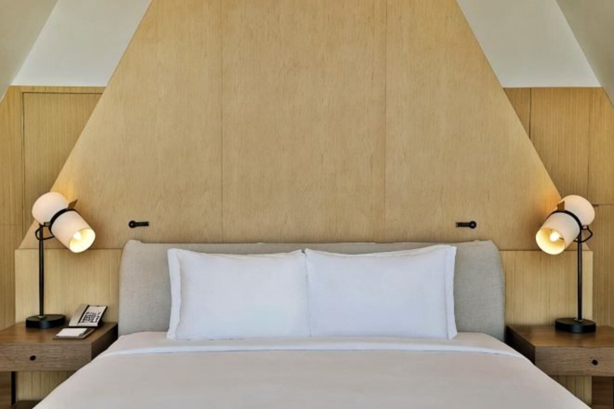 A hotel room with a wooden headboard, a bed with white bedding, two bedside lamps, and a nightstand with a phone.