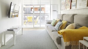 Ein modernes Wohnzimmer mit einer Couch, einem kleinen Tisch, einer Bank und einem an der Wand befestigten Fernseher. Ein Schiebetürenfenster führt in einen Außenbereich mit einem gelben Stuhl.