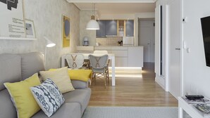 Ein modernes Wohnzimmer mit grauem Sofa, gelben und gemusterten Kissen, einer Essecke mit weißem Tisch und Stühlen sowie einer Küche mit grauen Schränken und einer Pendelleuchte.