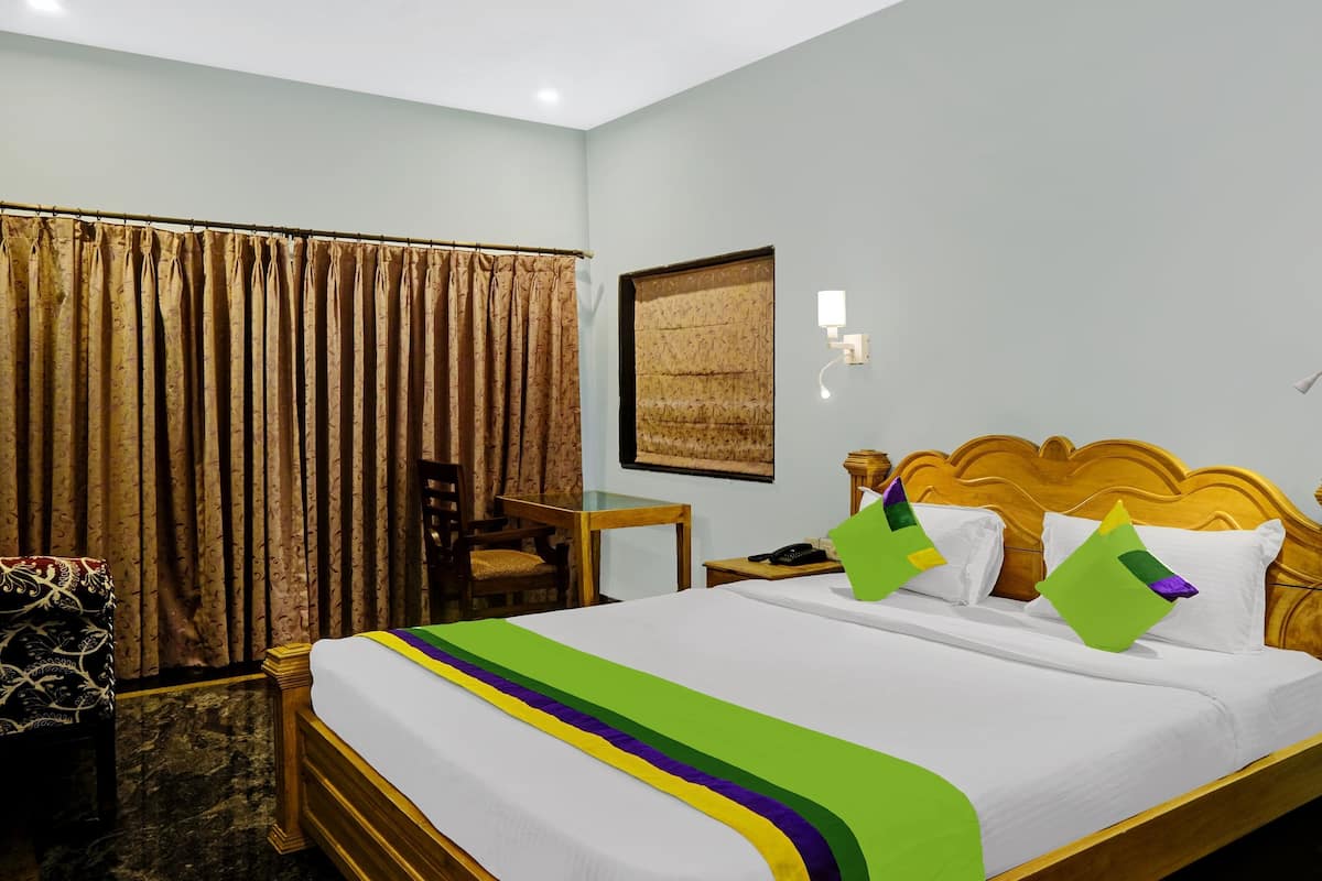 A hotel room with a bed, a chair, a table, curtains, and a wall-mounted lamp.