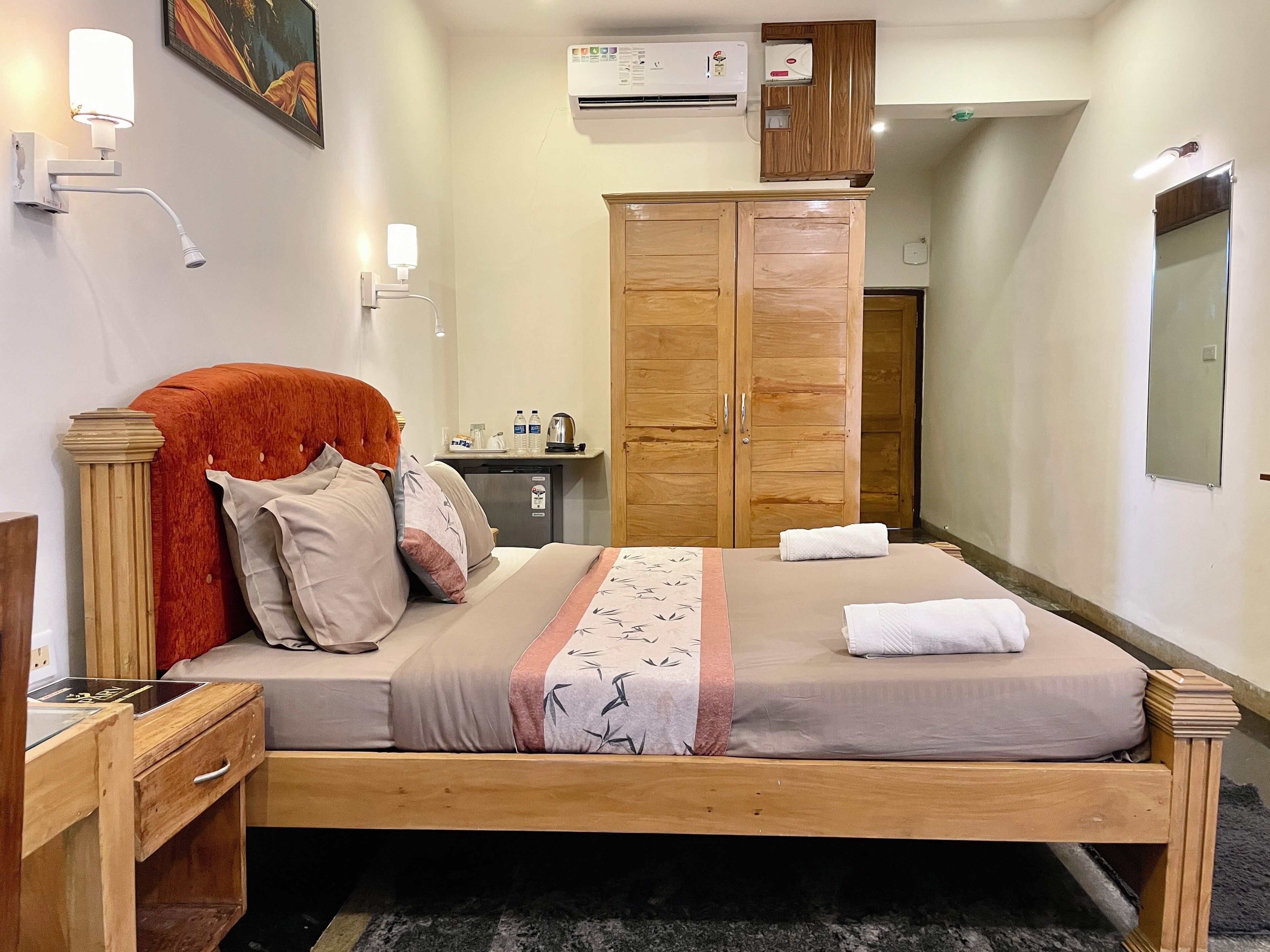 A neatly arranged hotel room with a bed, a wooden wardrobe, a small desk with a lamp, and an air conditioning unit.