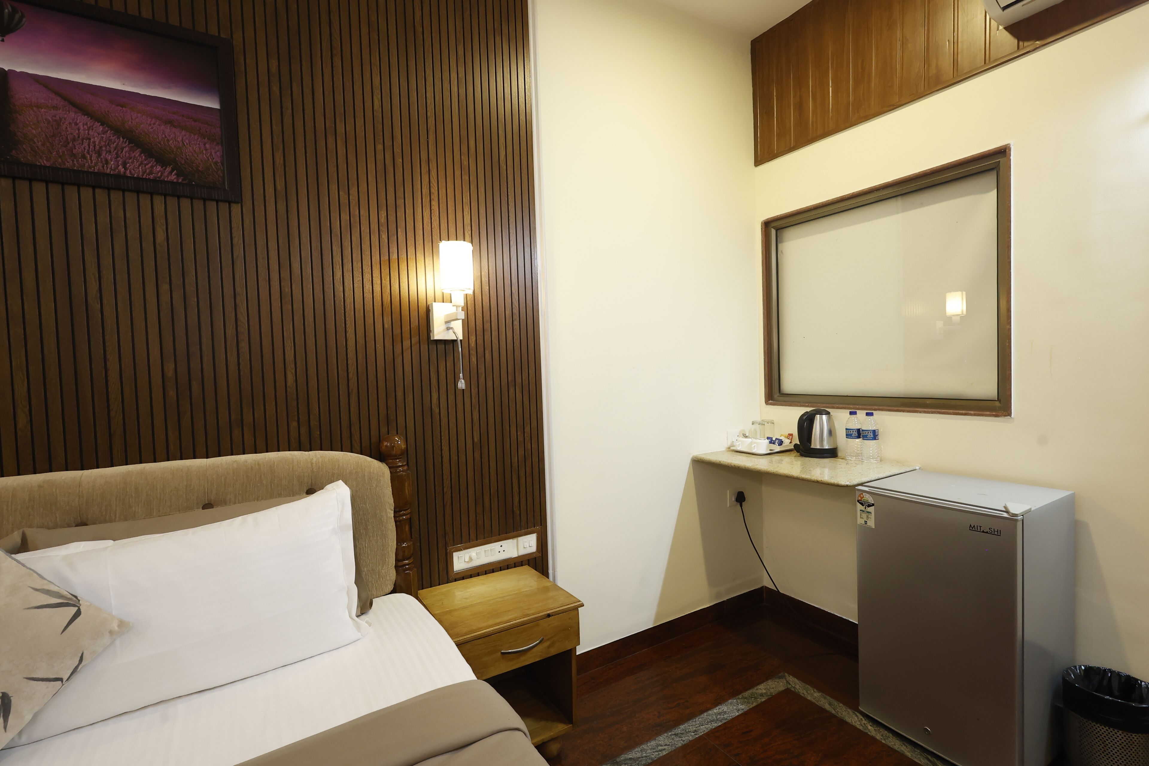 A hotel room with a bed, a nightstand, a desk with a computer, a refrigerator, and a wall-mounted mirror.