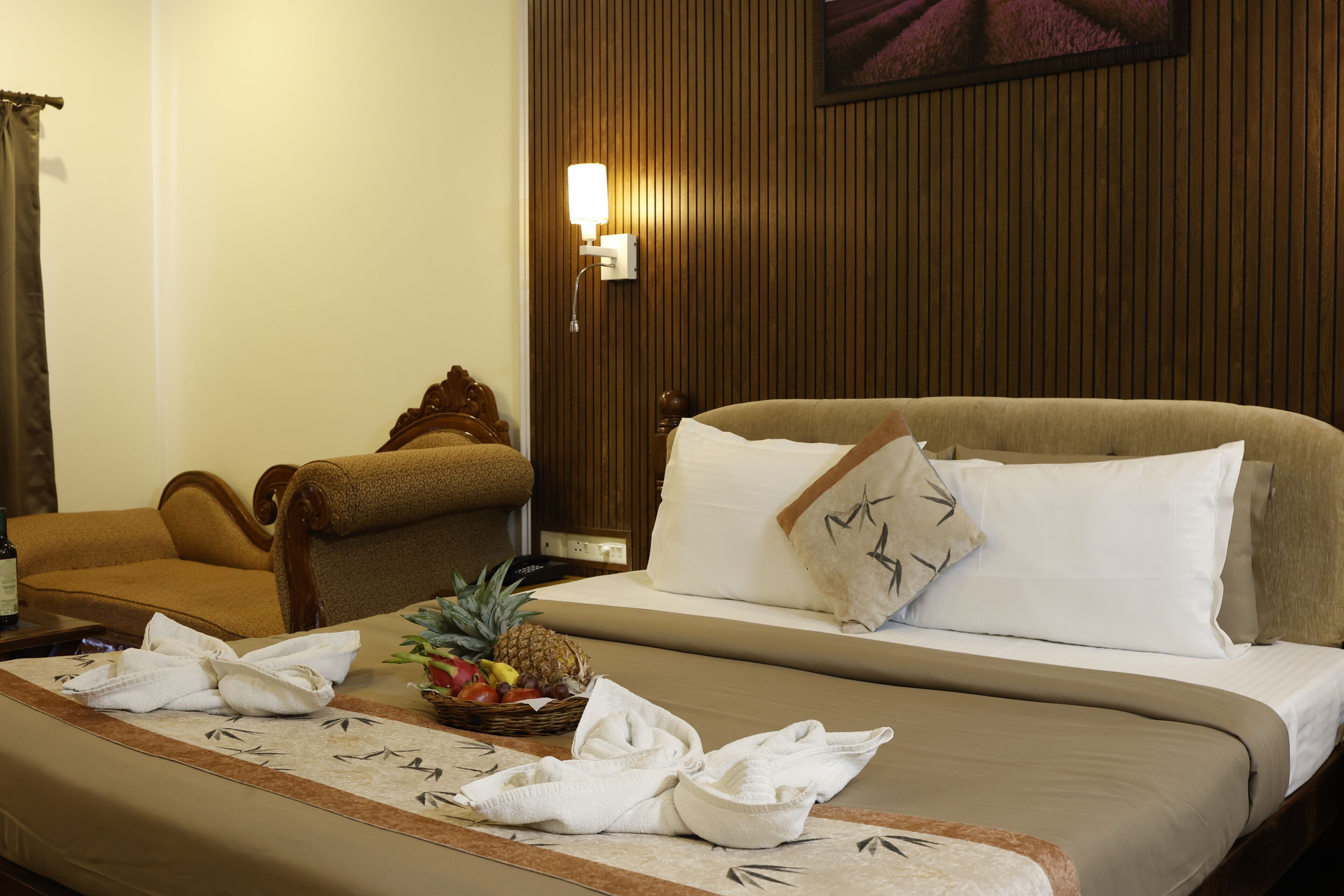 A hotel room with a bed, a wooden headboard, a nightstand with a lamp, a chair, and a fruit basket on the bed.