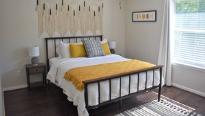 Um quarto com cama, mesas de cabeceira, ventilador de teto, um painel de macramé, uma moldura com imagem e uma janela com persianas.
