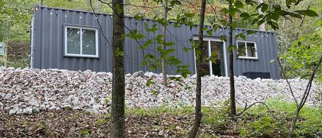 Une cabane moderne dotée d’une grande fenêtre et de fondations en pierre, entourée d’une forêt où les feuilles tombées recouvrent le sol.