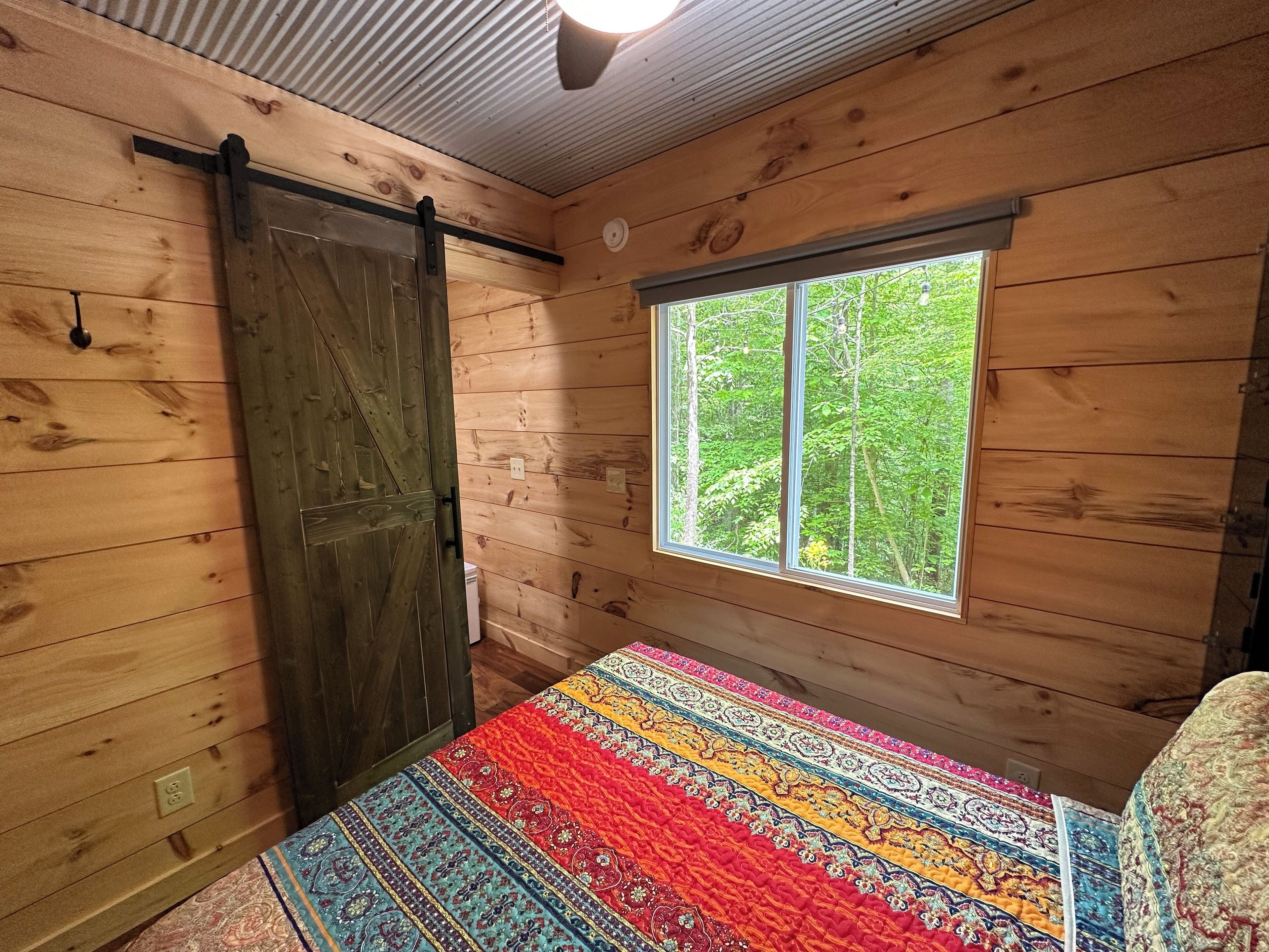 Ein Zimmer einer Holzhütte mit großem Fenster, einer Schiebetür und einer farbenfrohen Steudecke auf dem Bett.