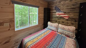 Quarto de cabana de madeira com uma janela grande, um ventilador de teto e uma cama com roupa de cama estampada.