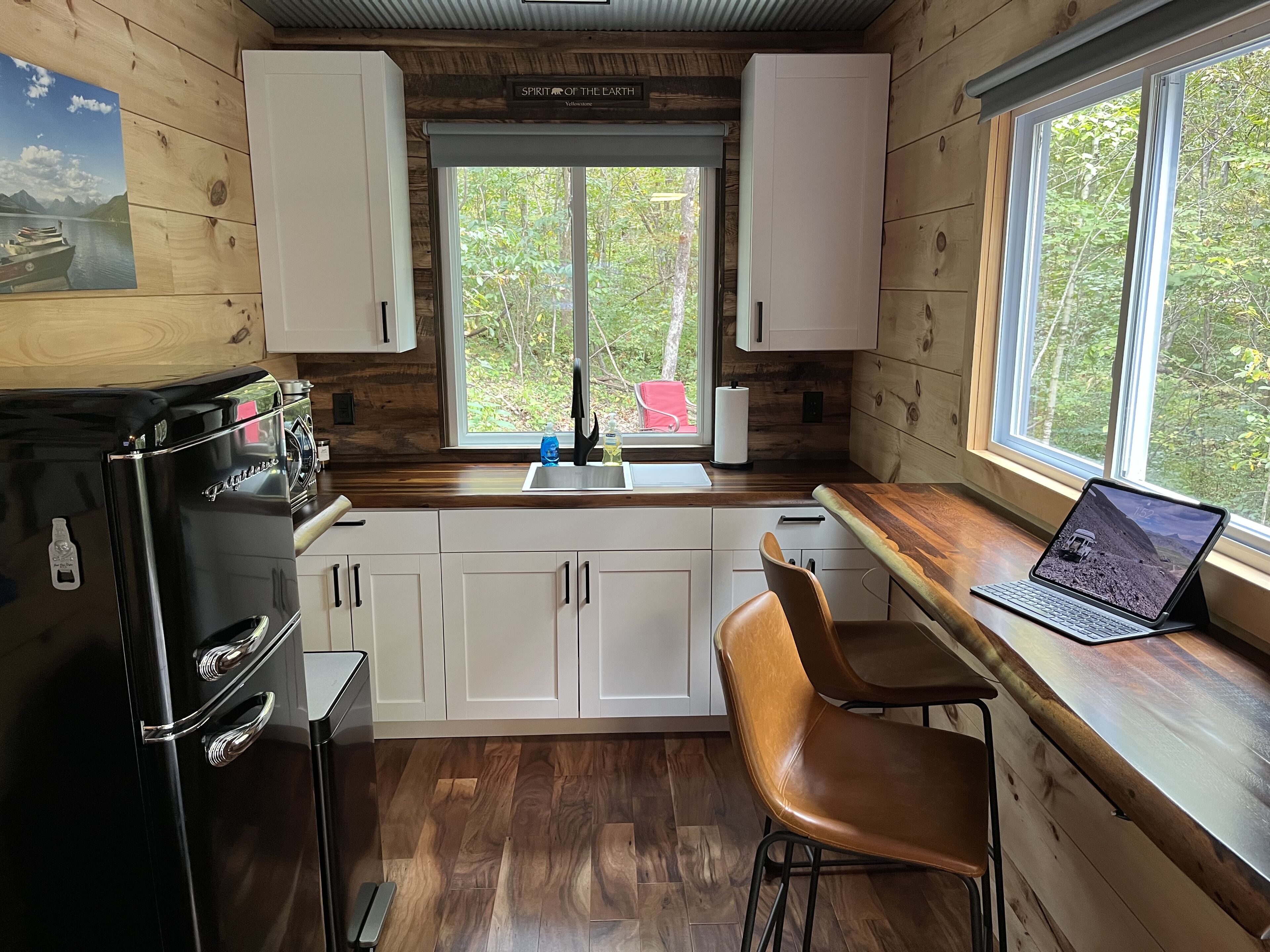 A compact kitchen with white cabinets, a black refrigerator, a sink, and a laptop on a wooden countertop.