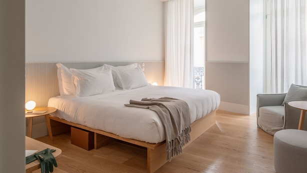 Un dormitorio moderno con una cama grande, una mesita de noche con lámpara, una silla y vistas al exterior a través de amplios ventanales.