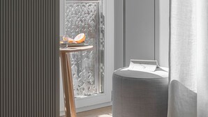 Una habitación moderna con una puerta de vidrio corredera que da a un balcón, una pequeña mesa de madera con un plato de fruta y un sillón tapizado en gris.