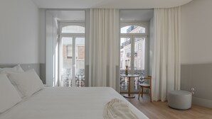 Una habitación de hotel moderna con una cama grande, una mesita redonda y vistas a la ciudad a través de amplios ventanales.