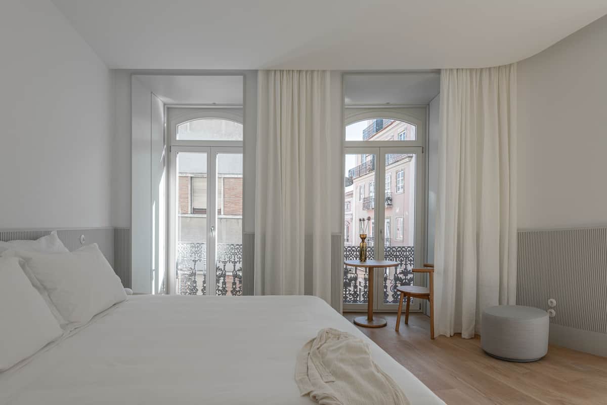 A modern hotel room with a large bed, a small round table, and a view of the city through large windows.
