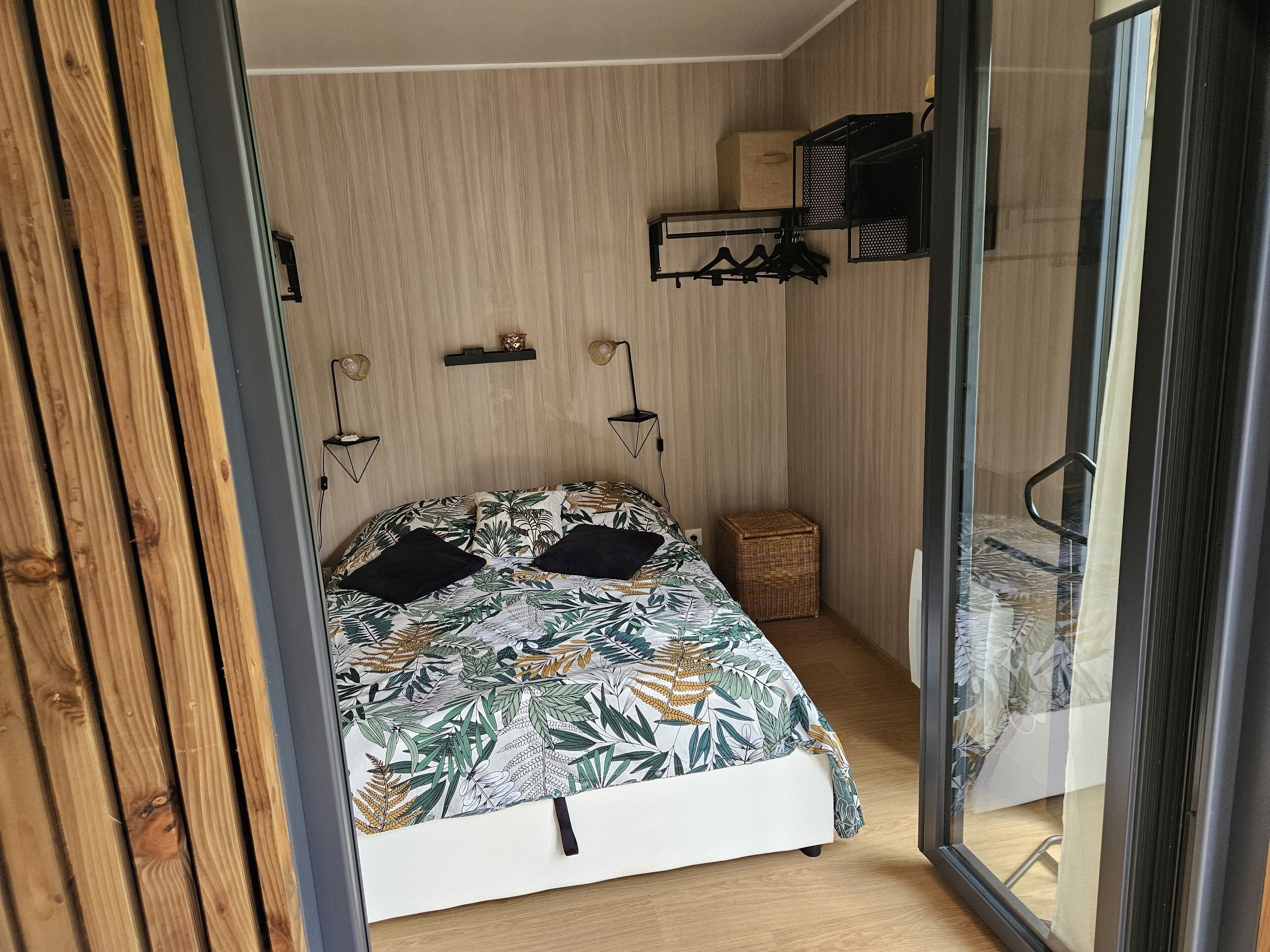 A bedroom with a bed, a small side table, a hanging shelf with clothes, and a window with a curtain.