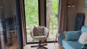 Una habitación con sofá, sillón, alfombra redonda y puerta de vidrio que da a un balcón con árboles.