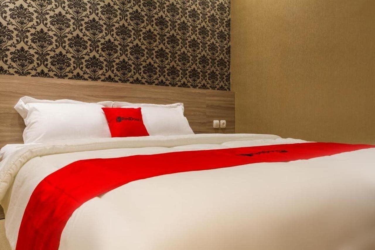 A neatly made bed with white and red bedding, a red pillow, and a patterned wallpaper in the background.