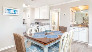 Un comedor con una mesa de madera, sillas de estampado azul y blanco, y una cocina al fondo.