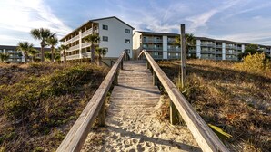 Una pasarela de madera que conduce a un edificio frente a la playa, con varios balcones y un sendero de arena.