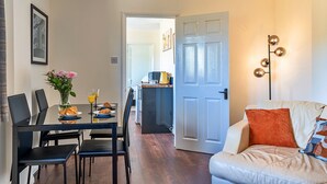 A dining area with a table set for two, a sofa with cushions, and a kitchen in the background. - The Annexe at Rosebud Cottage (Wedmore)