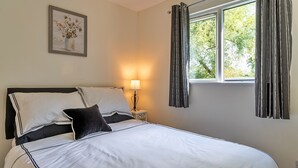 A neatly made bed with white bedding and black accents, a bedside lamp, a window with curtains, and a framed artwork on the wall. - The Annexe at Rosebud Cottage (Wedmore)