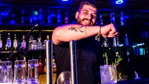 Ein Barkeeper mit Tätowierung am Arm, eine Bar mit Gläsern und Flaschen sowie ein Getränk mit Zitronenscheibe und Rosmarinzweig.