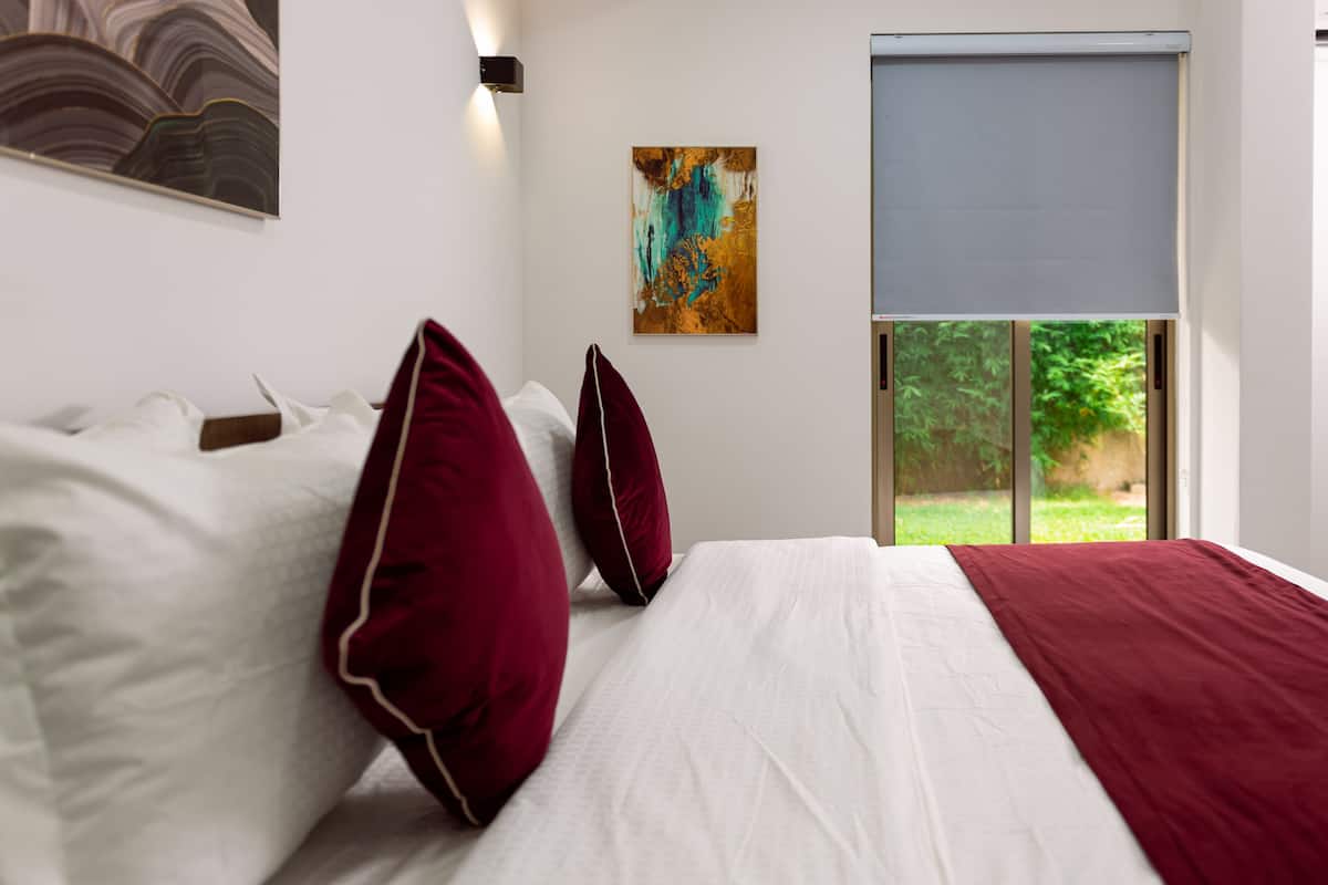 A modern bedroom with a large bed, white bedding, and red pillows. There is a painting on the wall and a window with a view of greenery.