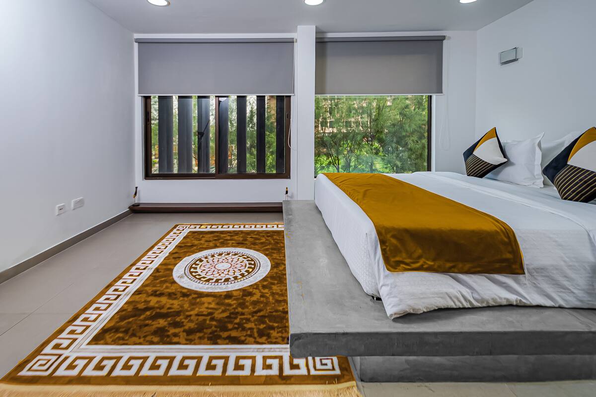 A modern hotel room with a large bed, a patterned rug, and a window with a view of greenery.