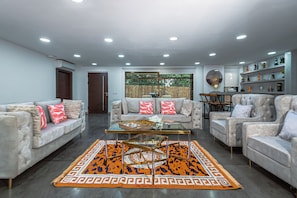 A modern living room with a glass coffee table, two sofas with patterned cushions, a patterned rug, and a bookshelf with various items.