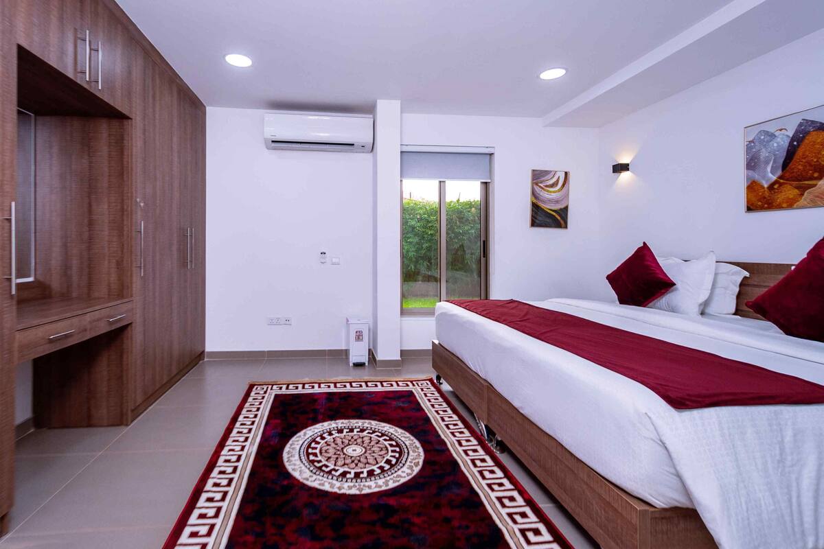 A modern bedroom with a large bed, a wooden wardrobe, an air conditioning unit, and a view of greenery through a sliding door.