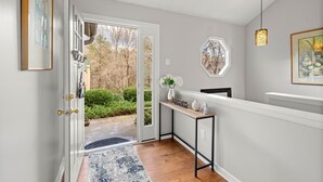 Un hall d’entrée moderne avec une porte vitrée donnant sur un jardin, une petite console ornée d’objets décoratifs et un luminaire suspendu.