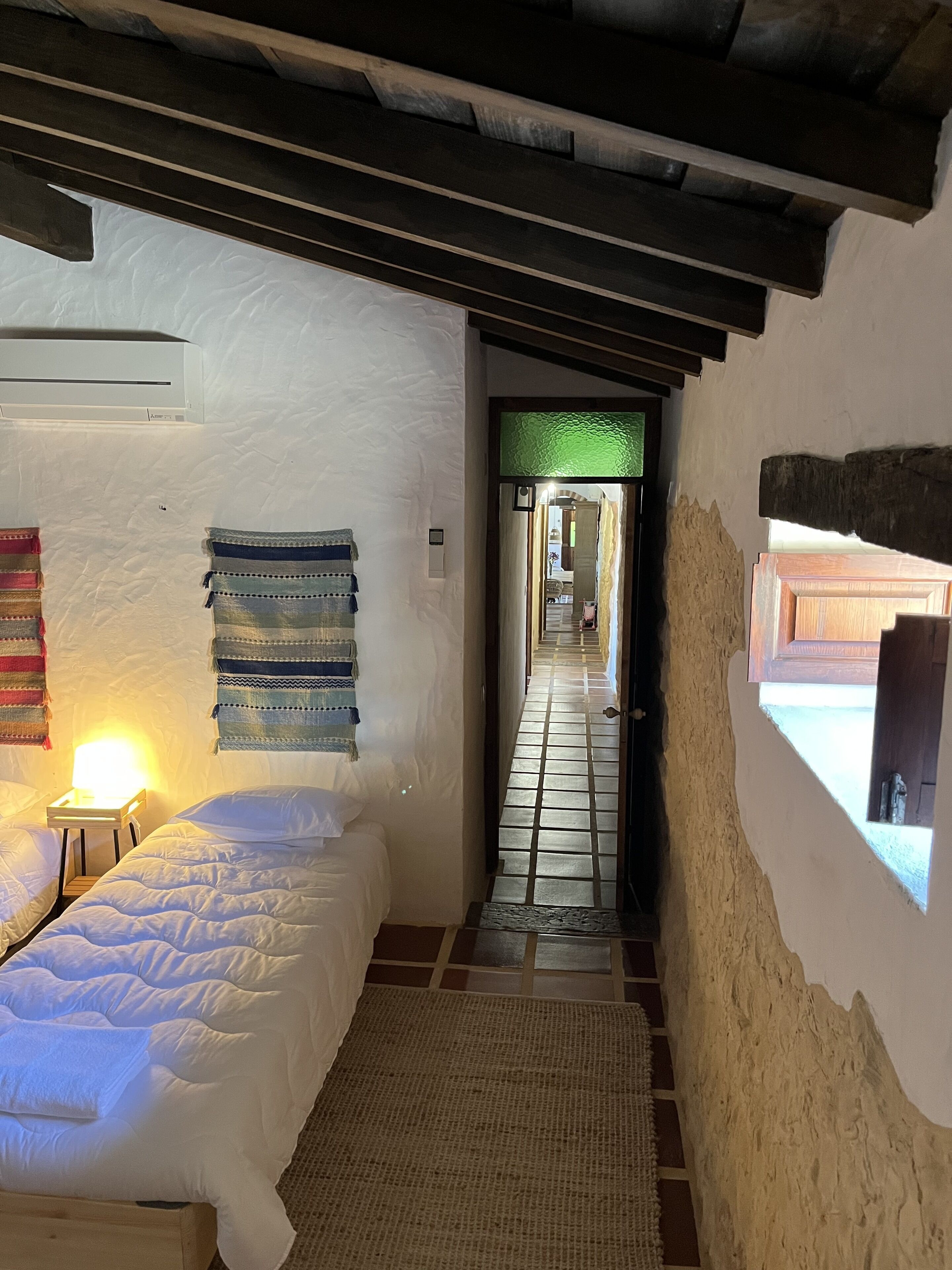 A narrow hallway with a bed on the left, a small table with a lamp, and a window on the right.