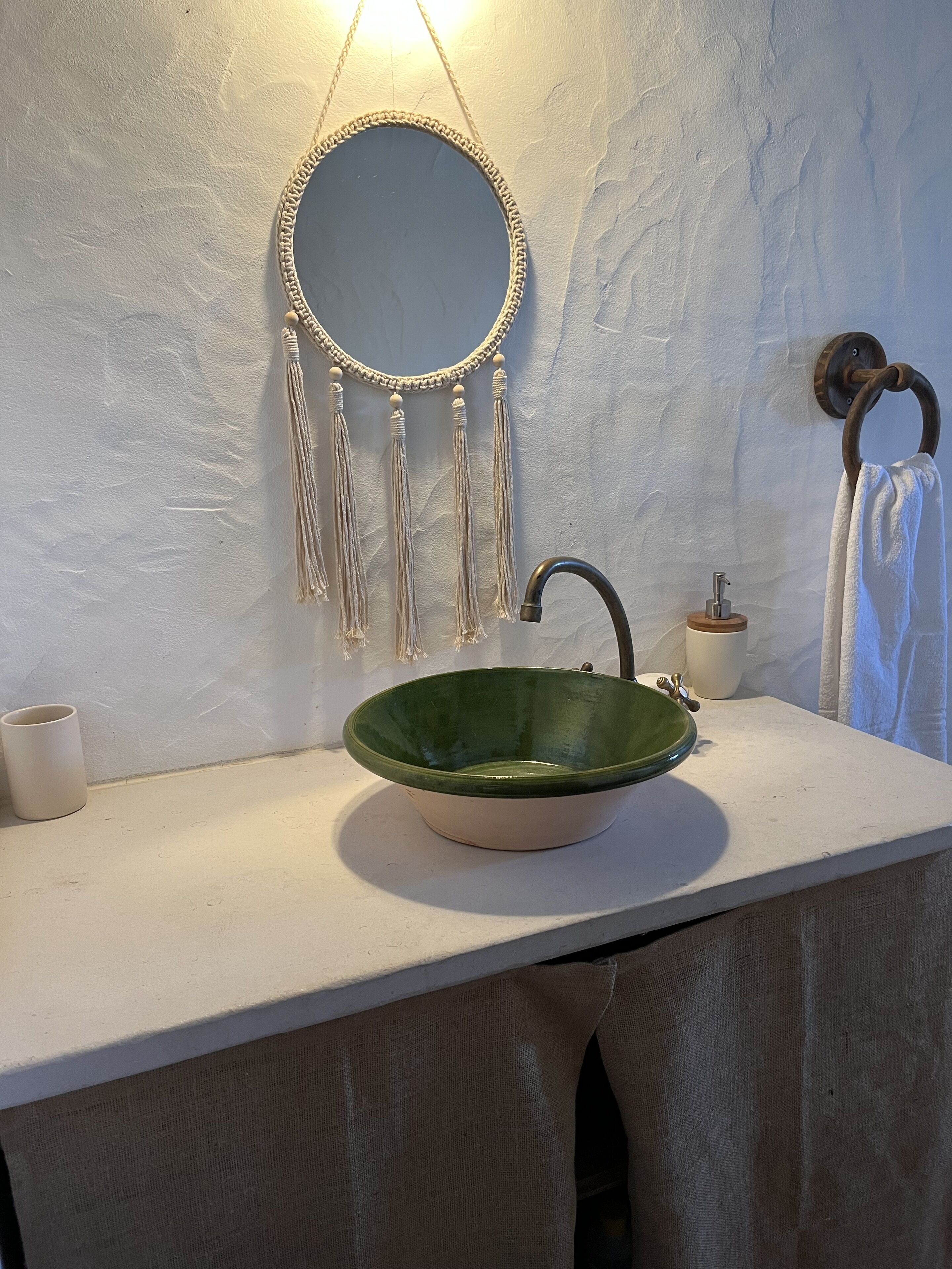 Un baño con un espejo redondo, un lavamanos verde, un perchero para toallas y una taza.