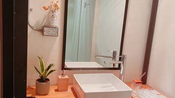 Une salle de bain avec un lavabo blanc, un miroir, une plante en pot, une petite boîte en bois et un panier en osier.