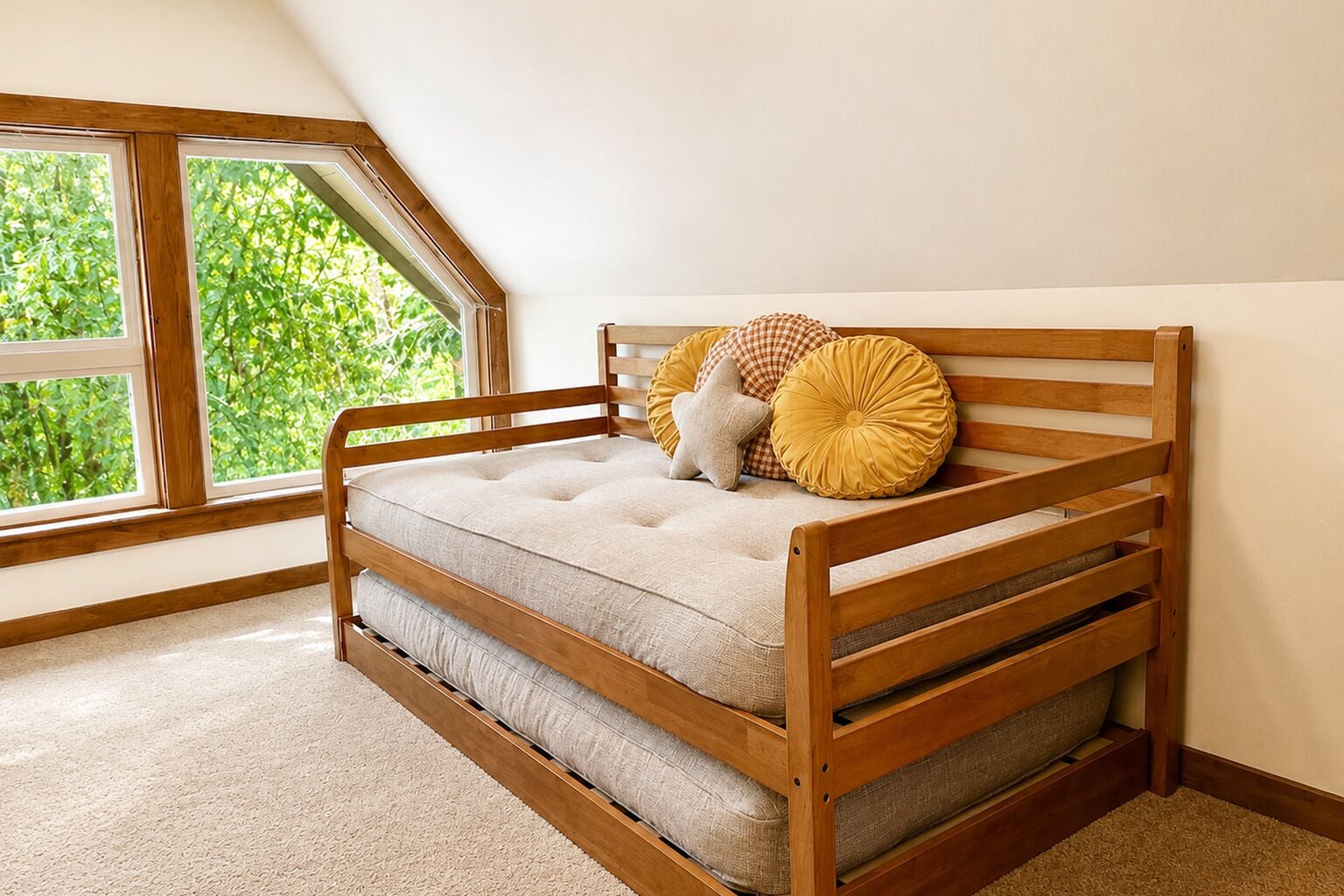 Ein Holzbett mit Etagen, ausgestattet mit einer gepolsterten Matratze, einem sternförmigen Kissen und einem runden Kissen in einem Zimmer mit geneigter Decke und einem Fenster mit Blick auf Grünflächen.