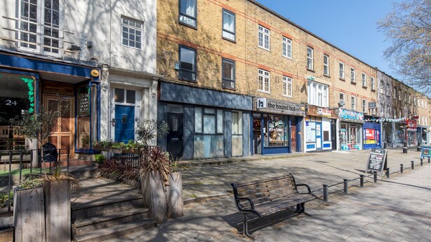 A street with a row of shops, including a bakery and a cafe. There are steps leading up to the shops, and a bench is visible on the sidewalk.